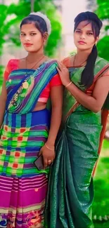 Two women in colorful saris, standing outdoors with vibrant green background.