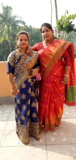 Two women in vibrant sarees with a natural background.