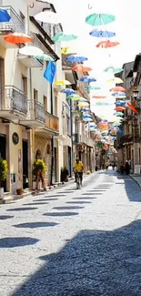 Street with colorful umbrellas suspended above, casting shadows on cobblestones.