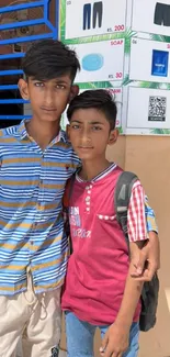 Two young individuals in colorful attire stand outdoors with a blue patterned background.