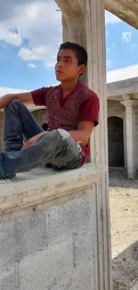 Young person in casual attire sitting in unfinished building under blue sky.