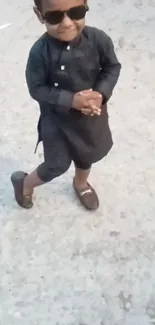 Child in black outfit with sunglasses on a gray pavement.