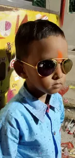 Young boy in blue shirt and sunglasses with bright yellow background.