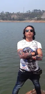 Cool tattooed man in sunglasses poses confidently by a scenic lake.