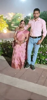 A couple standing in a well-lit park at night with a floral backdrop.