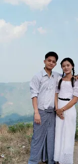 A couple in traditional attire poses with a scenic mountain view and clear sky.