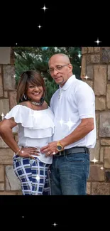 Smiling couple in casual attire with stone wall background.