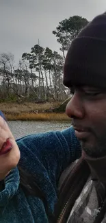 Couple embracing by a calm lake with forest backdrop.