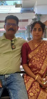 Couple seated indoors, woman in saree and man in casual shirt.