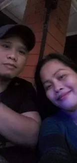 Couple in a cozy evening setting, smiling toward the camera with warm lighting.