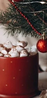 Red mug of hot cocoa with marshmallows beside Christmas tree decorations.