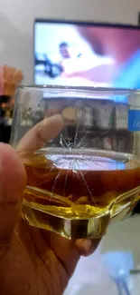 Hand holding cracked whiskey glass with TV background.