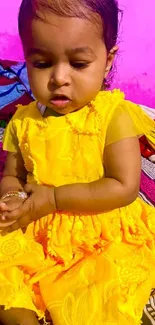 Cute baby in a vibrant yellow dress sitting on a colorful bedspread.