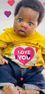 Cute baby in yellow outfit with pink heart saying 'Love You' on wallpaper.