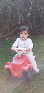 Cute baby riding a red toy car in a garden.