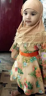 Young girl in floral dress and beige hijab, standing indoors.