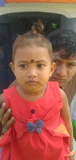 A cute child wearing a red dress with a bow, standing with an adult.