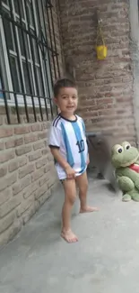 Young boy in soccer jersey with plush frog toy outside.