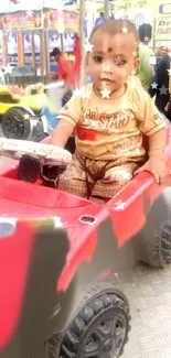 A toddler enjoys a ride in a red toy car at a carnival.