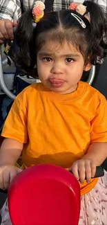 Little girl in orange shirt riding a red scooter outdoors.