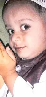 Cute child with hands in prayer wearing a white cap and brown outfit.