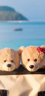 Two teddy bears on a beach with a blue ocean backdrop.