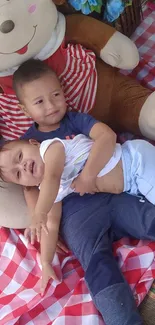 Two toddlers enjoy a picnic with a large teddy bear on a checkered blanket.