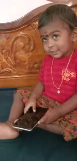 Cute toddler holding a smartphone in a red shirt, sitting on a bed.