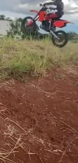 Motorcycle jumps over grassy hill, dirt backdrop.