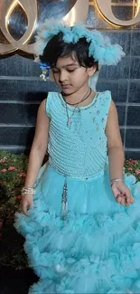 Child in an elegant blue dress standing against a floral and wooden backdrop.