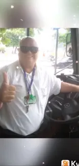 Smiling bus driver gives thumbs up in bright outdoor setting.