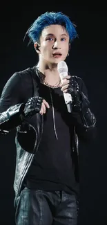 Blue-haired performer with microphone on stage, wearing gloves and black outfit.