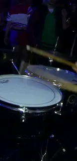 Close-up of drumsticks hitting drums in a dark, lively scene.