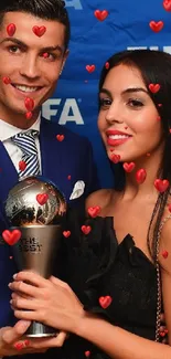 Elegant couple at award ceremony holding a trophy with blue backdrop.