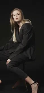 Elegant person in a black suit against a dark background, seated stylishly.