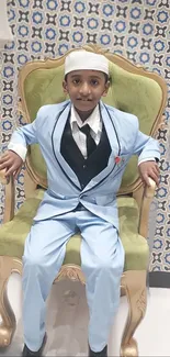 Boy in light blue suit sitting on a green chair against a tile-patterned wall.