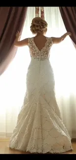 Bride in lace gown next to bright curtain.