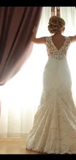 Elegant bride in lace gown by window with rich brown curtains.