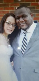 Elegant couple in formal attire against a brick wall, smiling joyfully.