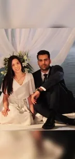 Couple in elegant attire sitting together with a white draped background.