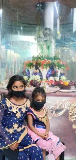 Two children in masks before an ornate floral setting.