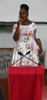 Woman in a floral dress speaking on stage with a red podium.