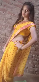 Woman in yellow saree against rustic brick wall.