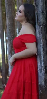 Woman in elegant red dress leaning against tree in forest.