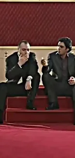 Two men in suits seated on red carpet steps, elegant setting.