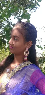 Woman in a blue and purple sari with elegant jewelry and a green leafy background.