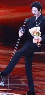 Person in elegant black suit holding flowers on stage.