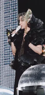 Stage performer in black attire with floral accents and a microphone.