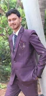 Man in a burgundy suit leaning against a pole with lush green background.