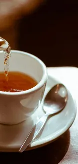 Close-up of tea being poured into cup with saucer and spoon in warm light.